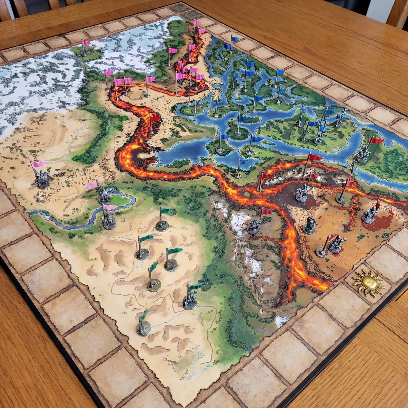 The Kaladorn main board on a wooden table, with flag-bearing miniatures in pink, blue, green and red spread across deserts, lakes and a glowing lava river, and a golden sun token on the border track.